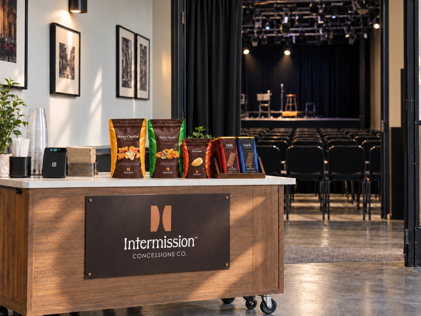 A portable Intermission concessions cart with a marble countertop holding Honey Cheddar, Spicy Guac and Salted Peanuts snack mixes and milk and dark chocolate bars, its mounted Intermission Concessions Co. sign standing before an empty black-box theater
